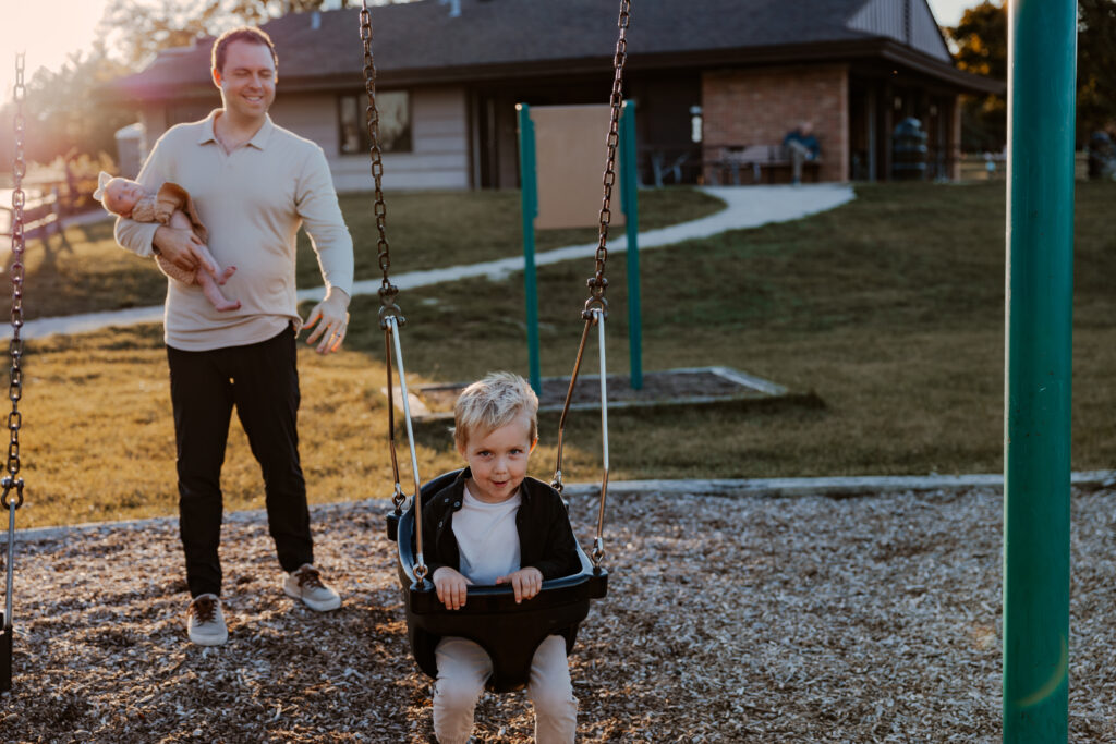 Dad pushing his little boy on a swing while gently holding a newborn at family friendly park in Chicago, enjoying a family moment outdoors.