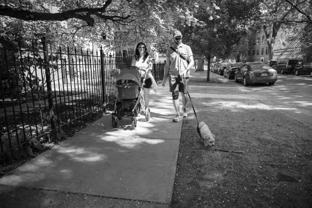 Postpartum mom and husband pushing a stroller along the sidewalk in family friendly park in chicago.